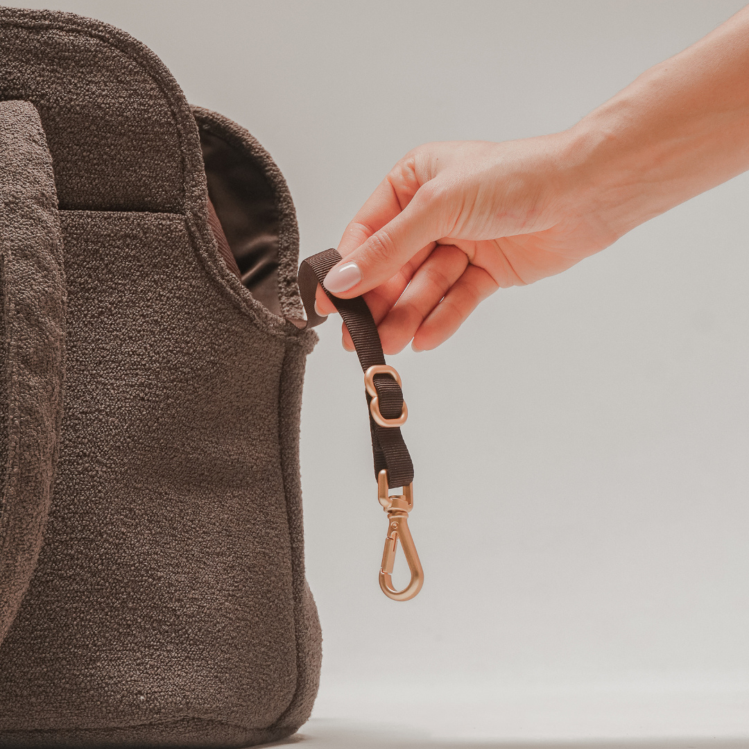 Hand attaching a brown leather strap to a brown bag on a light gray background