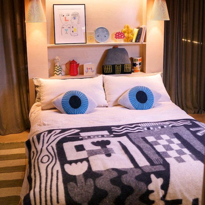 A cozy bedroom scene featuring a bed with white bedding and two distinctive round blue and black eye-shaped pillows. A black and white geometric patterned blanket is draped over the foot of the bed, set against a warm beige headboard with built-in shelves holding decorative items, framed art, and a small basket.