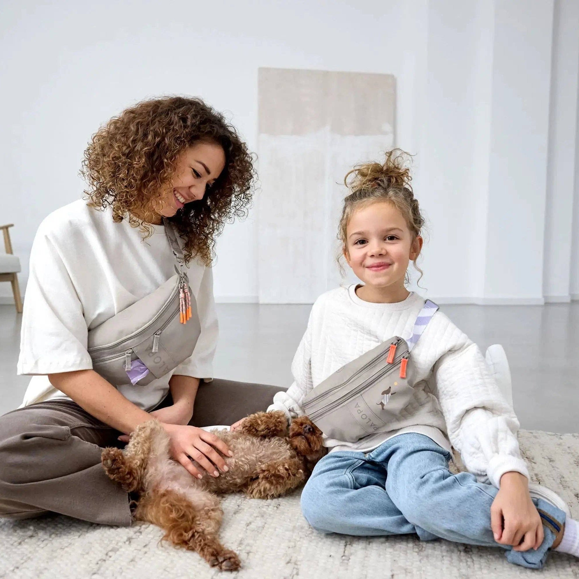 Woman and child sitting on a rug with a small dog, both wearing baby carriers.