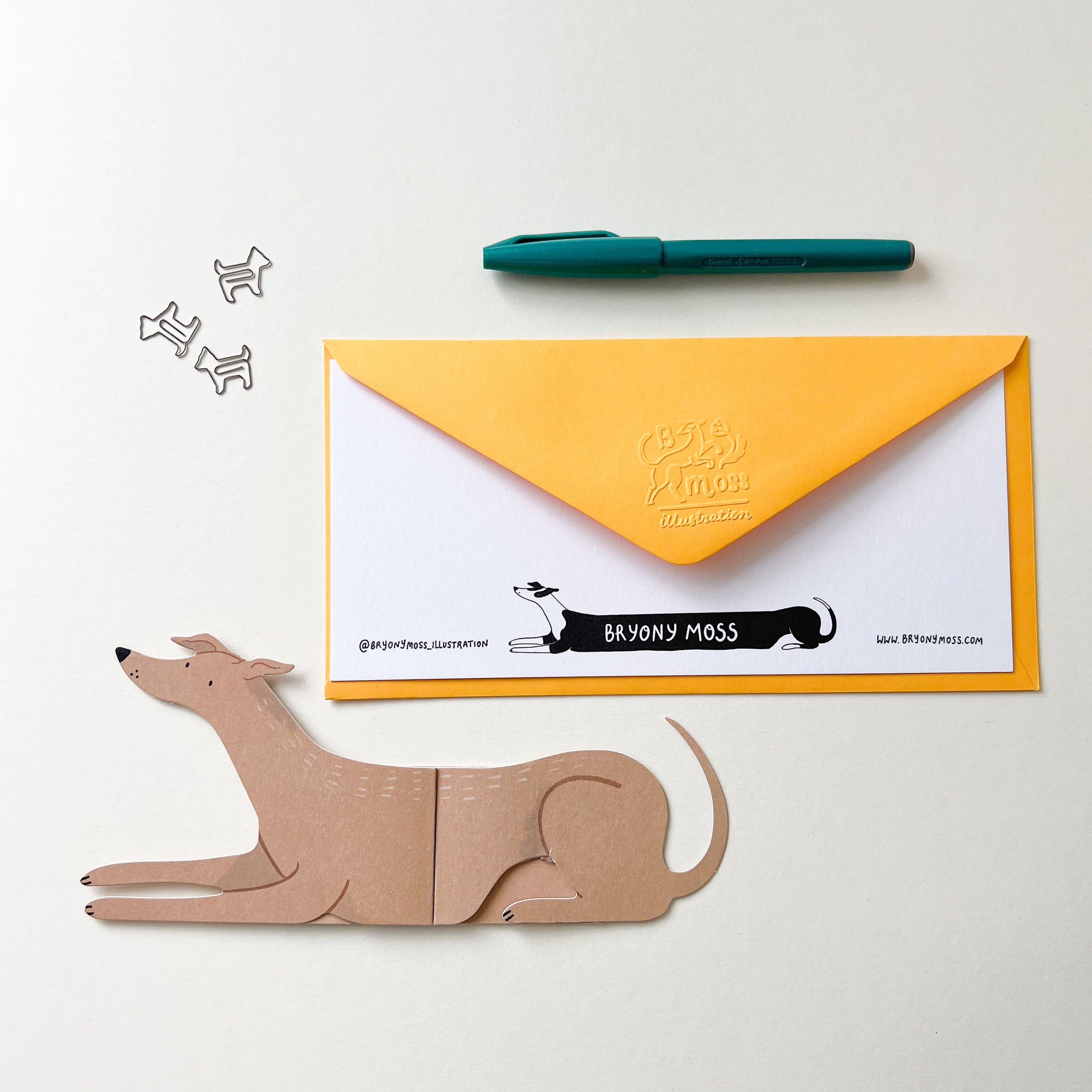 Dog-shaped paper clip and yellow envelope with Bryony Moss branding on a white background