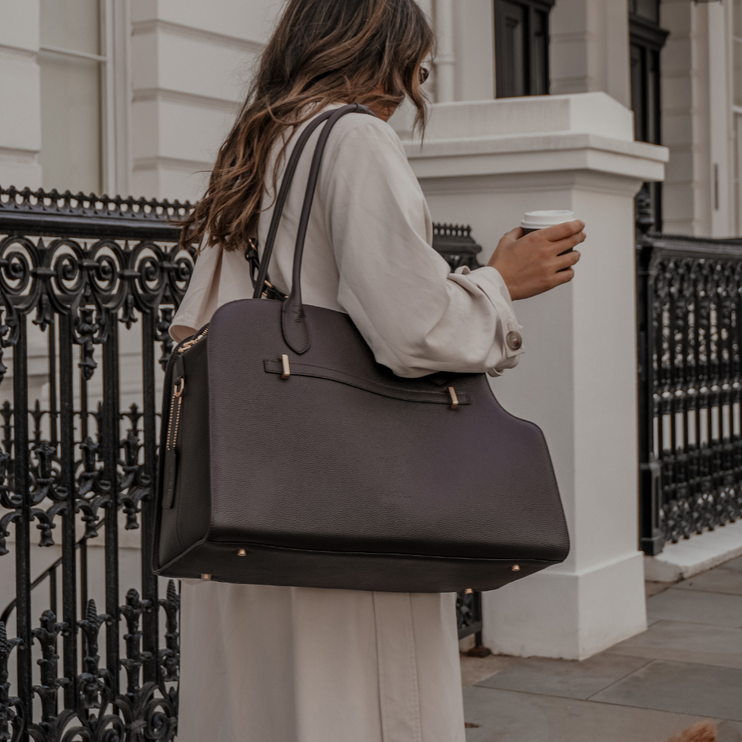 Woman holding a large brown leather bag and a coffee cup, walking a dog on a city street.