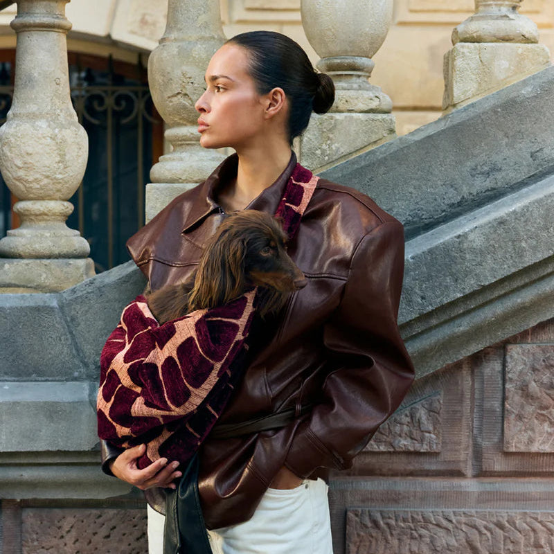A woman wearing an oversized dark brown leather jacket and white pants stands on stone steps, holding a small brown dog wrapped in a burgundy and beige patterned blanket.
