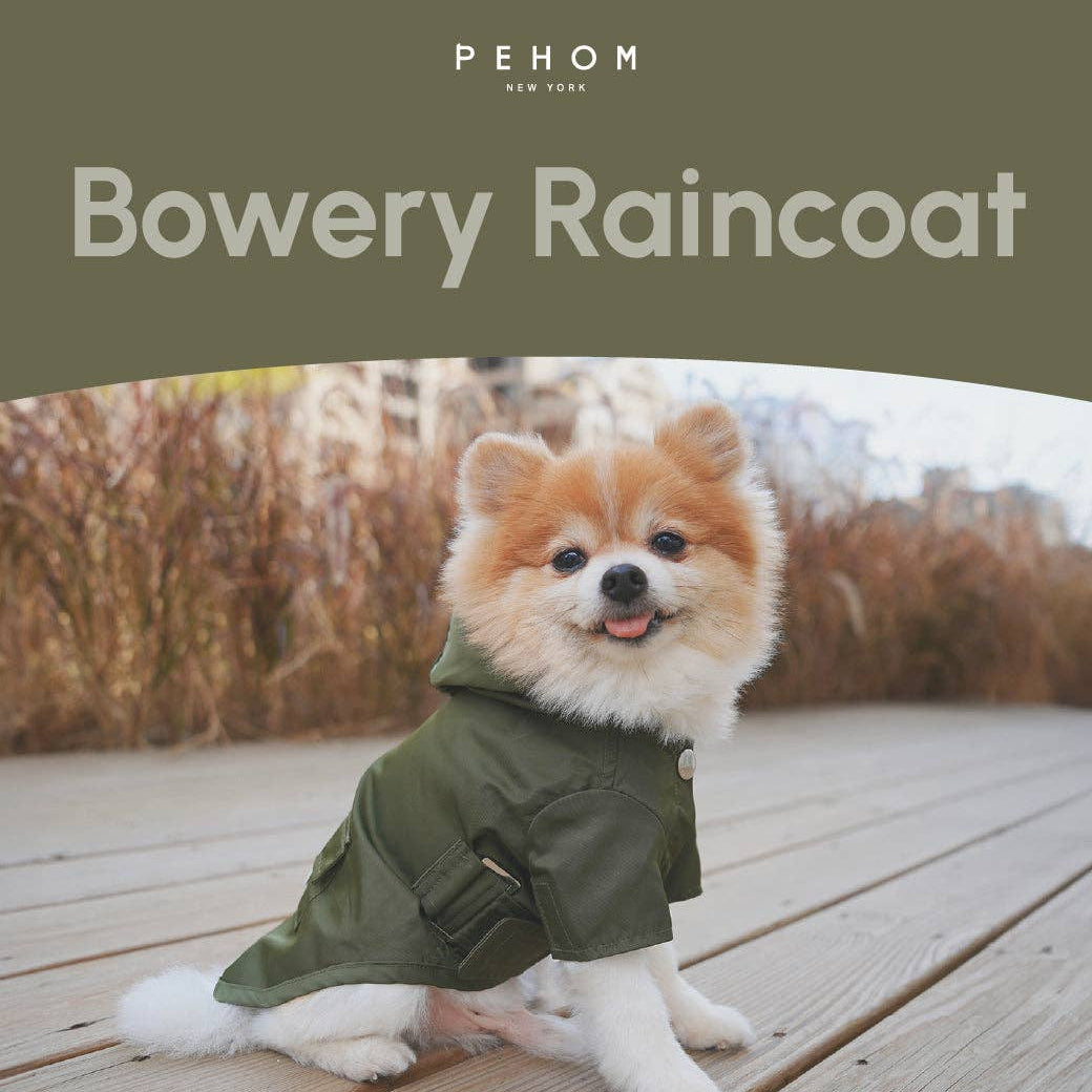 A small fluffy dog wearing an olive green hooded raincoat sits on a wooden boardwalk outdoors. The top of the image has a dark olive banner with the text 'PEHOM NEW YORK' and 'Bowery Raincoat'.
