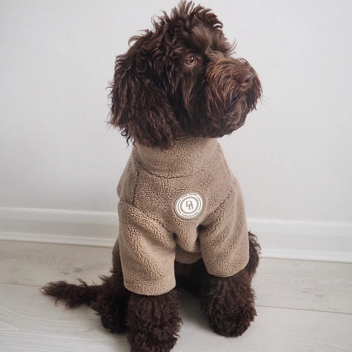 Dog wearing a beige fleece onesie with a brand logo on a white floor.