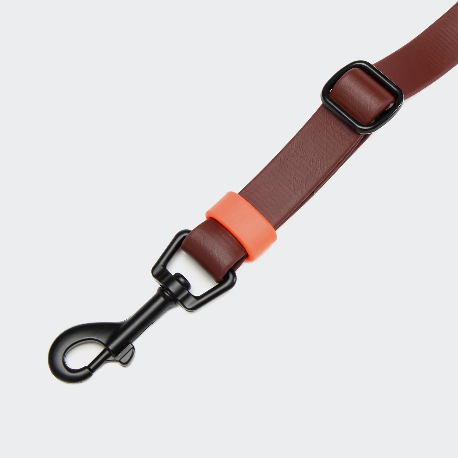 Brown leather dog leash with black clip and orange accent on a light gray background