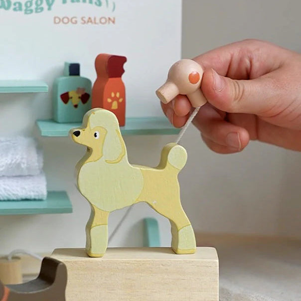 Wooden dog toy on a stand with a hand pulling a string, set against a background of 'Waggy Tails Dog Salon'.