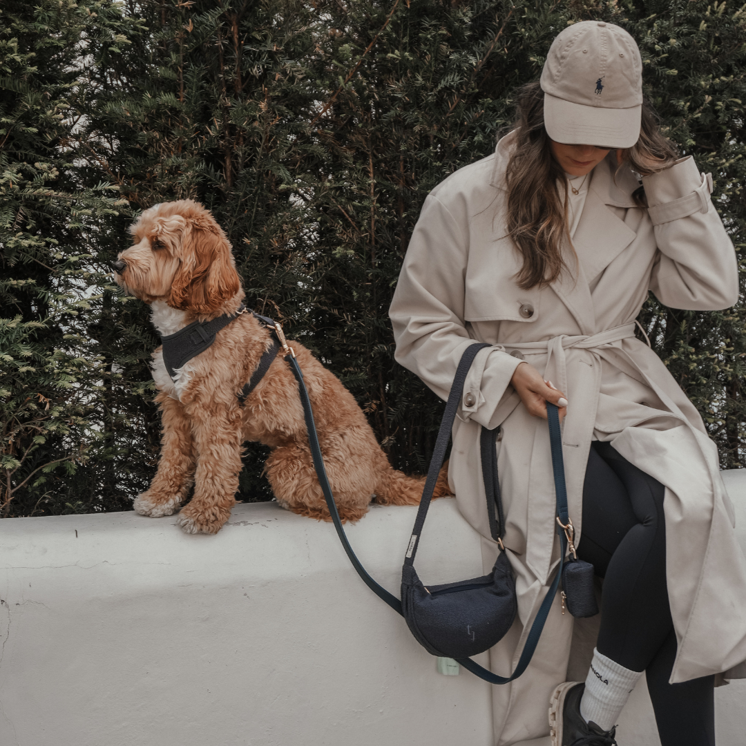 Woman in a beige coat and cap with a dog on a leash, standing on a white wall with greenery in the background.
