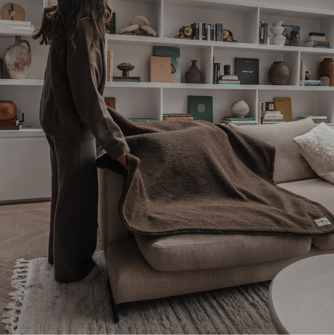 Woman arranging a brown blanket on a beige sofa in a living room with bookshelves.