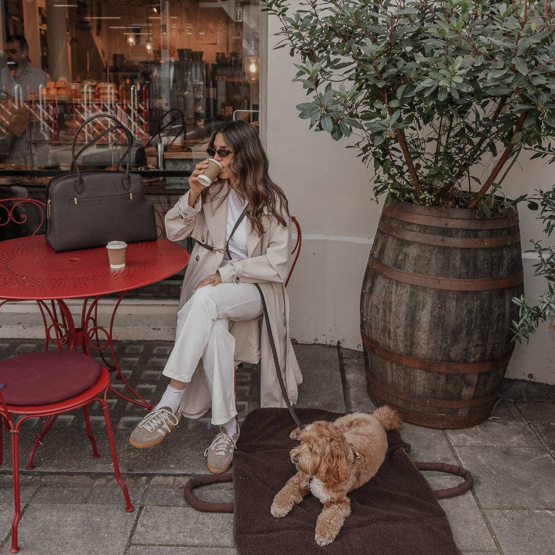 Woman sitting on a sidewalk with a dog on a mat, drinking from a cup.