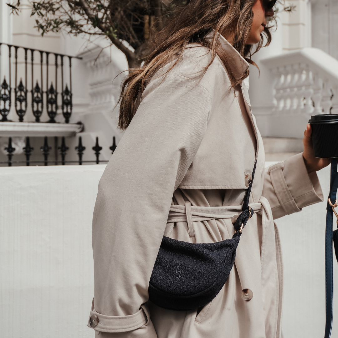 Woman in a beige trench coat with a black bag and hat, standing outdoors.