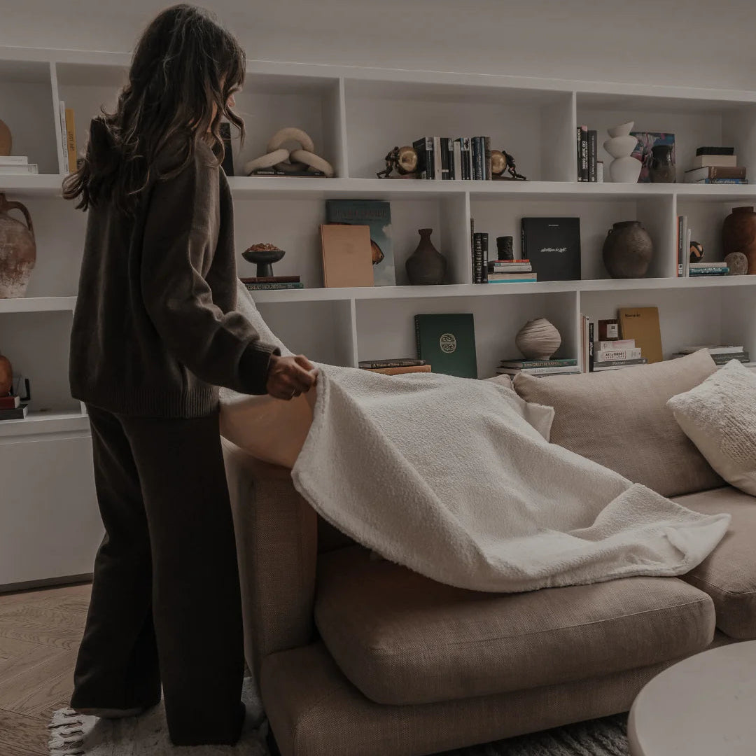 Person helping another person onto a couch in a living room with bookshelves in the background.