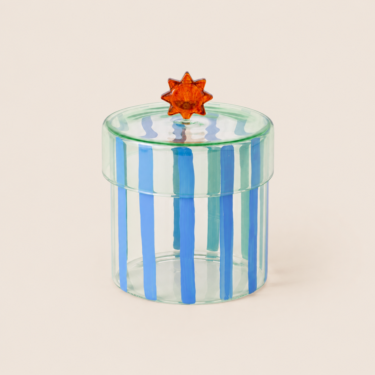 A small cylindrical clear glass box with vertical blue stripes and a translucent orange star-shaped lid knob, photographed on a plain warm beige background.