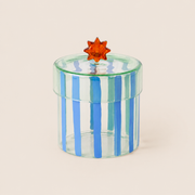 A small cylindrical clear glass box with vertical blue stripes and a translucent orange star-shaped lid knob, photographed on a plain warm beige background.