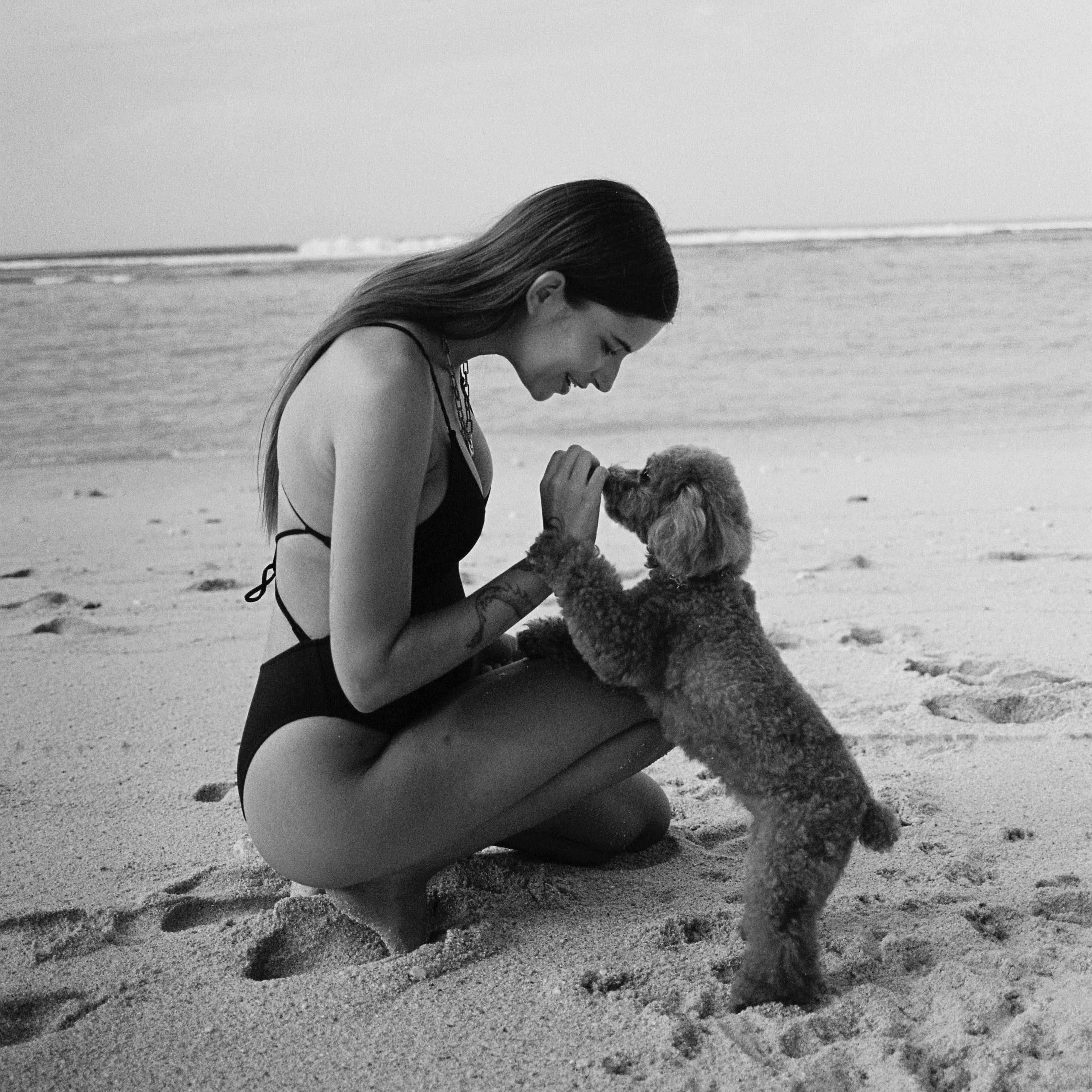 Woman in swimsuit kneeling on beach with small dog