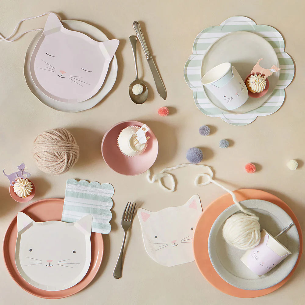 Cat-themed paper plates, cups, and cutlery on a beige surface with yarn balls and small desserts.